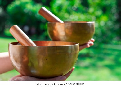 Two Singing Tibetan Bowls In Female Hands On A Background Of Green Lawn. Singing Bowls For Healing In Female Hands. Occupation Of Alternative Tibetan Indian Medicine. Sound Therapy.