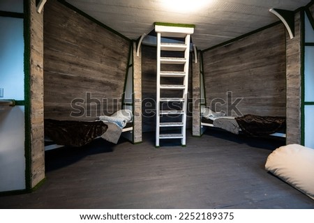Two single beds with linen mounted in niches of a room. Pyramid shape bungalow summer house. Angled surrounded walls, white stairs, gray horizontal wooden wall planks. Nonstandard frame construction.