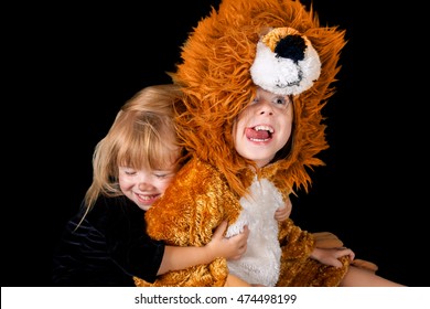 Two Sisters In Costumes Being Cute And Silly.  A Little, Blond Cat Hugs Her Older Sister The Lion.  The Lion Has Her Tongue Out And Is Making A Funny Face.