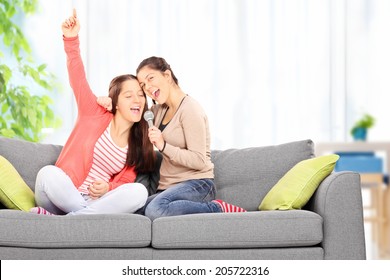 Two sisters having fun singing on microphone seated on a sofa at home