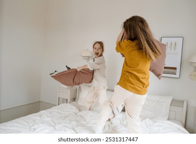 Two sisters are having a playful pillow fight on a bed, laughing and having a great time. The image captures the fun and innocence of childhood and sibling bonding. - Powered by Shutterstock