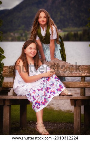 Two sisters with long hair in summer dress and dirndl on a bench in front of a lake
