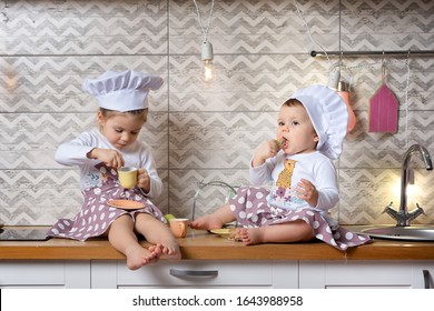 Two sisters one-year-old and three-years-old girls in a cook hats sit on the kitchen table and drink tea with cookies