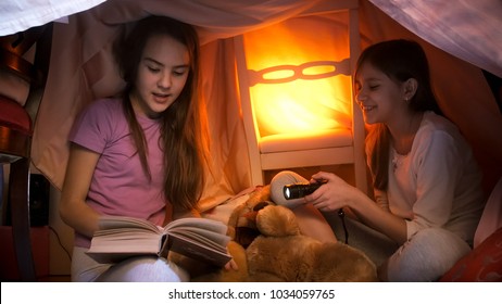 Two sisters in pajamas reading book in tent made of blankets at home