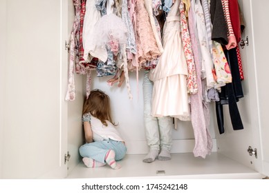 Two Sisters Play Hide And Seek Inside A Closet With Lots Of Colorful Flowered Dresses. One Of The Girls Reaches Out To Touch Her Sister, Who Is Hiding In The Corner.