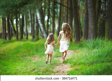 
Two Sisters Run Through The Forest  Holding Hands. Summer Sunny Day And Girls In Light Dresses. Beautiful Long Hair In A Child.