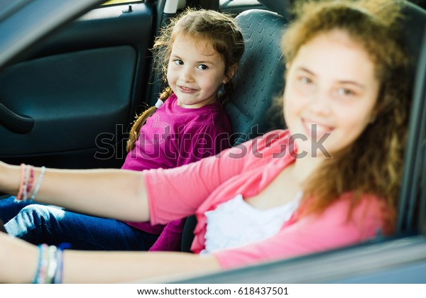 Two sisters are smiling to\
the camera while sitting in the family car.The eldest one is\
blurred.