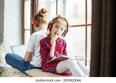 Two Sisters Study At Home. The Girl Writes In A Notebook. Children With Headphones Listening To An Audiobook. Teenager With Laptop. Big Window. Cozy Home.