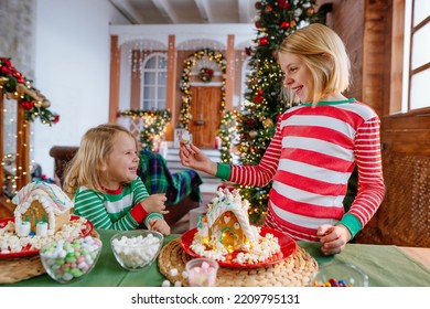 Two Sisters Wearing Striped Pajamas Decorating Tradition Christmas Cakes Gingerbread Houses With Icing And Candies At The Kitchen. Selective Focus.