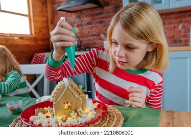 Two Sisters Wearing Striped Pajamas Decorating Tradition Christmas Cakes Gingerbread Houses With Icing And Candies At The Kitchen. Selective Focus.