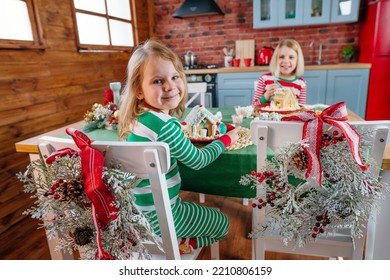 Two Sisters Wearing Striped Pajamas Decorating Tradition Christmas Cakes Gingerbread Houses With Icing And Candies At The Kitchen. Selective Focus.