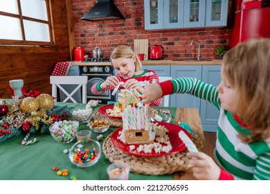 Two Sisters Wearing Striped Pajamas Decorating Tradition Christmas Cakes Gingerbread Houses With Icing And Candies At The Kitchen. Selective Focus.