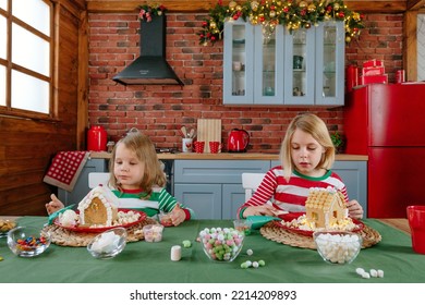 Two Sisters Wearing Striped Pajamas Decorating Tradition Christmas Cakes Gingerbread Houses With Icing And Candies At The Kitchen. Selective Focus.