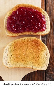 Two slices of toasted white bread, one with jam, the other without, on a cutting board, on old wooden background, top view