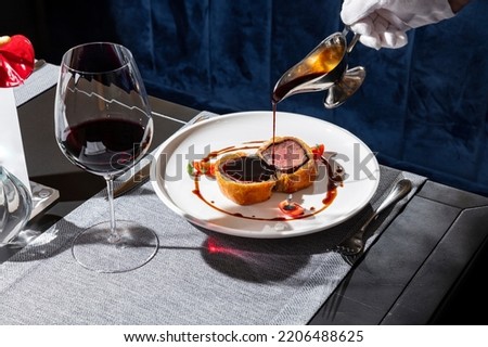 Two slices of wellington beef with tomatoes, sauce, herbs in a plate on the table with a napkin, sauce in a saucepan with cutlery and a glass of wine