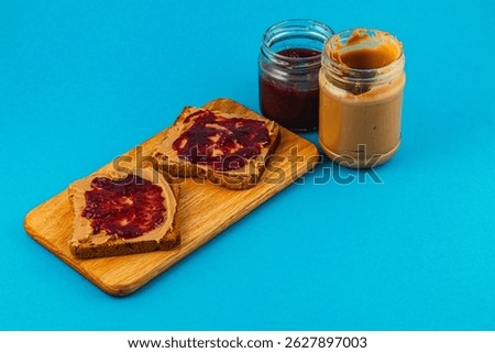 Two slices of whole grain bread topped with creamy peanut butter and grape jelly sit on a wooden cutting board alongside jars of spread.