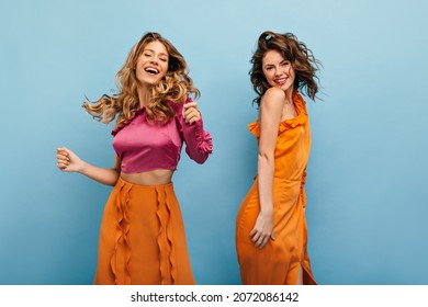 Two slim caucasian young girls vigorously posing on blue background. Beauties with wavy hair smile cute. Blonde in pink top and long skirt. Brunette in orange dress with bare shoulder.