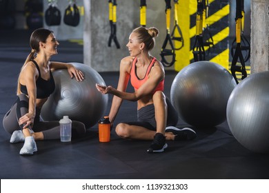 Two Slim Ladies Are Laughing During Conversation In Fitness Studio. They Are Sitting On Floor With Water Flacks Near Fitballs And Using Smartphone. Females Are Looking At Each Other While Relaxing