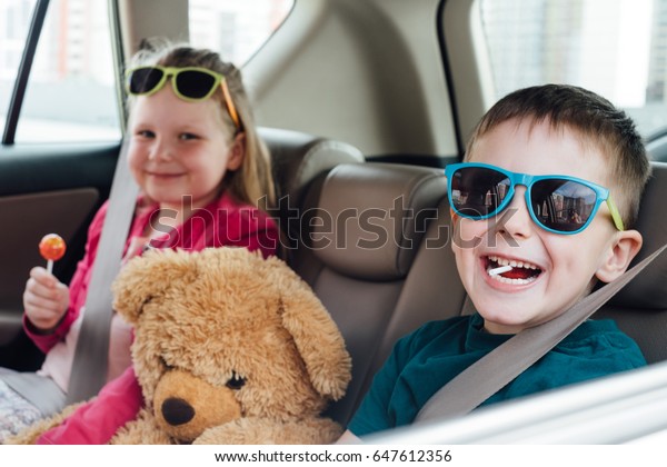 Two small beautiful\
children with toy bear in the car with candy and fasten seat belts.\
Brother and sister happy playing smiling. Children fastened with\
seat belts