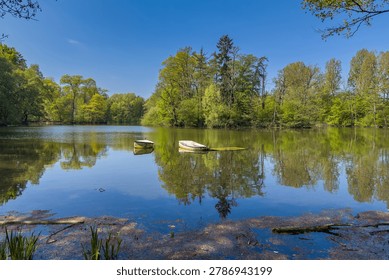 Two small boats float on a lake. Trees surround the water. The sky is blue with no clouds. It is a sunny day, and the scene reflects on the lake's surface. The area looks quiet and inviting.