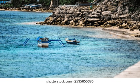 Two small boats are floating in the ocean near a rocky shore. The water is calm and blue, and the boats are blue and white - Powered by Shutterstock