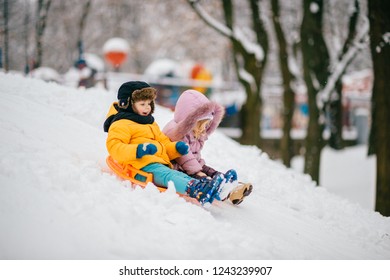 Two Small Cheerful Children Ride Board On Snow Hill On Clear Frosty Day. Brother And Sister Playing Outside In Winter. Sincere Children's Emotions In Snow