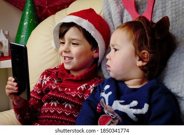 Two Small Children Are Sitting In A Room Decorated For Christmas, And Talking On A Video Link In The Phone With Their Relatives. One Boy In A Santa Hat, The Second Kid With Reindeer Horns On His Head.