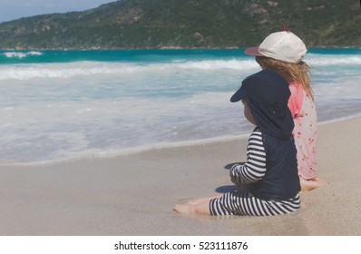 Two Small Children, Toddler Boy And Girl In Preschool Age Siblings Sitting In Sand Close To Ocean And Playing With Each Other, Wearing Sun Protective Swimming Clothing