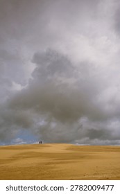 Two small figures venturing across an immense sand dune beneath a dramatic, cloudy sky