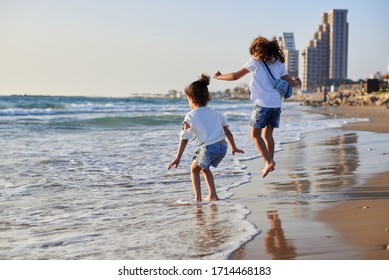 Two Small Girls Sisters, Playing Jumping On The Beach At Sunset. Beautiful Seaside Landscape Picture. Family Vacation At Tropical Resort In Summer.