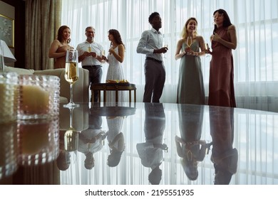 Two Small Groups Of Wedding Guests Or Members Of Bride Family With Champagne Standing In Front Of Table In Living Room At Party