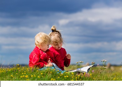 Two Small Kids In Preschool Age Reading Together. Siblings Bonding Outdoors. Big Sister Reading For Small Brother In Field. 