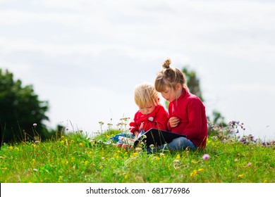 Two Small Kids In Preschool Age Reading Together. Siblings Bonding Outdoors. Big Sister Reading For Small Brother In Field. 