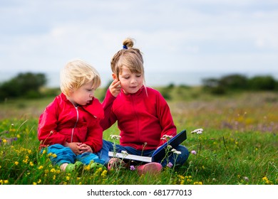 Two Small Kids In Preschool Age Reading Together. Siblings Bonding Outdoors. Big Sister Reading For Small Brother In Field. 