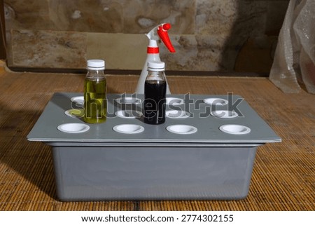 Two small plastic bottles containing yellow and dark purple liquid nutrients sit on a grey hydroponic tray with a white spray bottle behind it, arranged on a bamboo mat for indoor gardening.