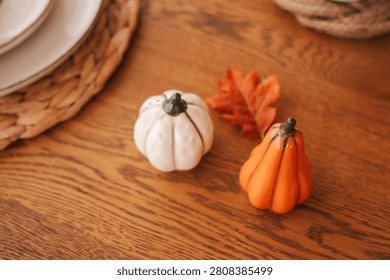 Two small pumpkins are placed on a wooden table. One pumpkin is orange and the other is white. Nearby there are some autumn leaves and plates arranged for decoration.