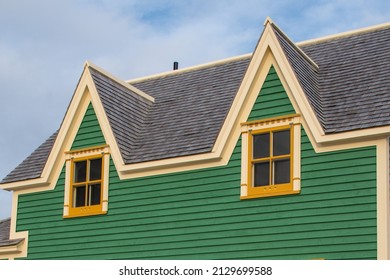 Two small single hung windows under gable or triangle shaped dormers on a vintage green exterior building wall with wood casement yellow trim. There's a blue sky with white clouds in the background