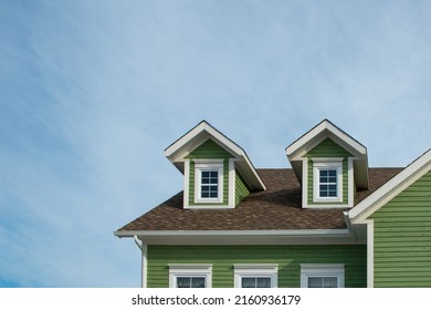 Two small single hung windows under gable or triangle shaped dormers on a vintage green exterior building wall with wood casement yellow trim. There's a blue sky with white clouds in the background