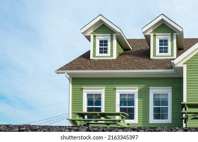 Two small single hung windows under gable or triangle shaped dormers on a vintage green exterior building wall with wood casement yellow trim. There are wooden picnic tables in front of the building.