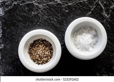 Two Small White Dishes Of Sea Salt Flakes And Freshly Ground Black Pepper. Top Down With Black Textured Slate Background