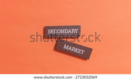Two small wooden rectangular blocks inscribed with the words secondary and market are positioned against a solid bright orange textured surface.