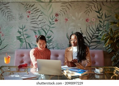 Two Smart College Girls Sit In A Coffee Shop With Books On A Table And Use A Laptop For An Online Class. Girls On Exchange Study In A Coffee Shop.