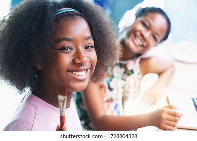 Two Smiling African Teen Girl Painting With Watercolour Paint In Room, Looking At Camera