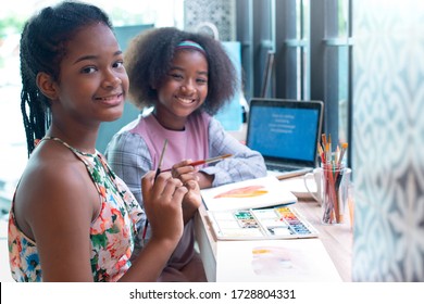 Two Smiling African Teen Girl Painting With Watercolour Paint In Room, Looking At Camera