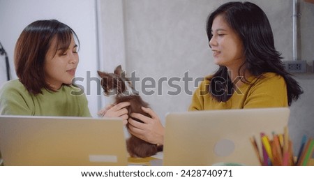 Two smiling Asian women enjoy a playful moment gaze affectionately at a cute Chihuahua between them sharing a tender moment in living room
