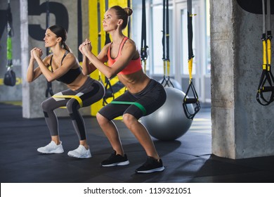 Two smiling athletic girls are squatting synchronously with outfit in fitness studio. Women are enjoying leg and buttocks work out with resistance band. Training together with friend in gym concept - Powered by Shutterstock