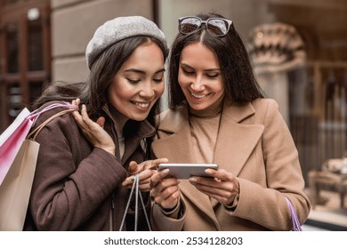 Two smiling attractive women girlfriends using holding smartphone looking on screen while shopping at the big city center urban street near magazine. - Powered by Shutterstock