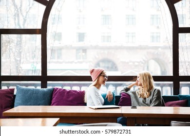 Two Smiling Beautiful Young Women Talking At The Table In Cafe