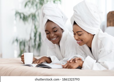 Two smiling black ladies lying on bed at home, reading magazine and drinking tea, copy space, closeup. Beautiful african american girlfriends having spa day at hotel, relaxing after beauty procedures - Powered by Shutterstock