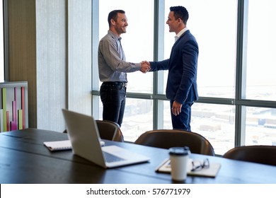 Two smiling businessmen shaking hands together while standing by windows in an office boardroom overlooking the city  - Shutterstock ID 576771979